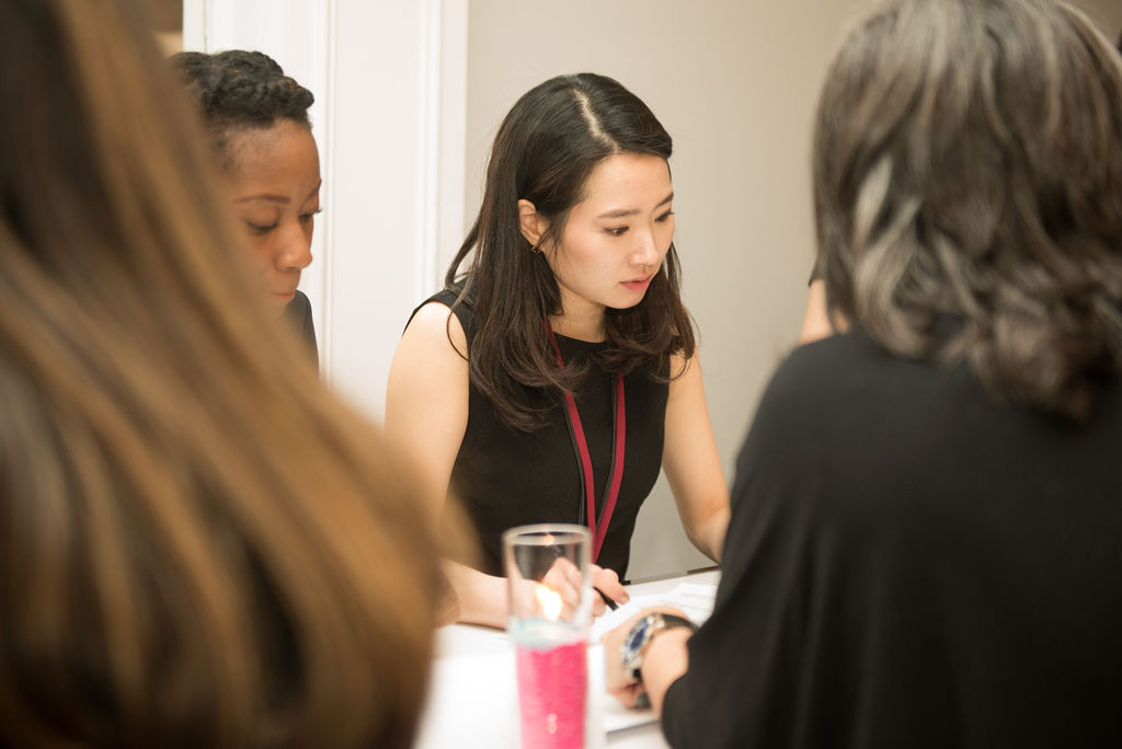 four women seated at a board room style education session. Two women have black tops seated back to the camera, the women facing the camera are Asian in a black sleeveless dress the other one is black and you cant see her clothes she is peeking over someones shoulder